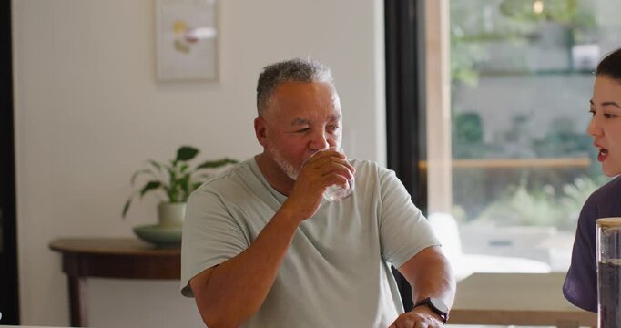 Senior African American man sipping from glass at kitchen island while Asian woman offering pitcher
