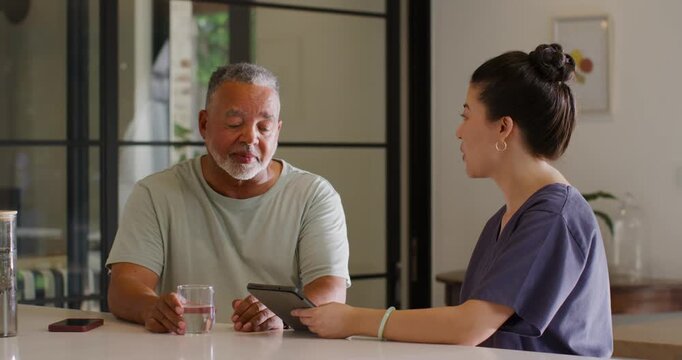 Diverse carer showing tablet demo at kitchen island, senior holding glass mask under chin sipping