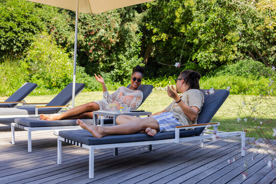 African American couple reclining and chatting on sun loungers on wooden deck with drinks, umbrella