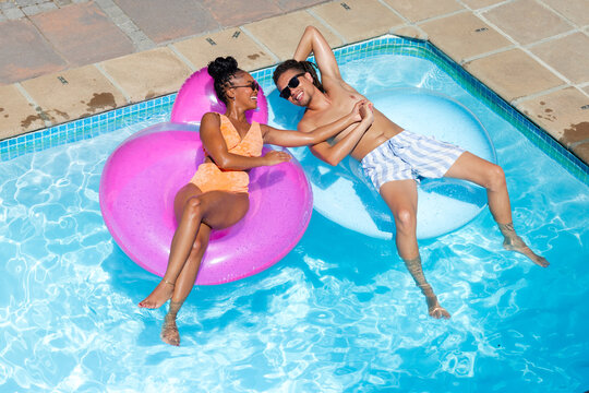 Couple reclining on pink and pale blue floats, holding hands, wearing sunglasses, floating in pool