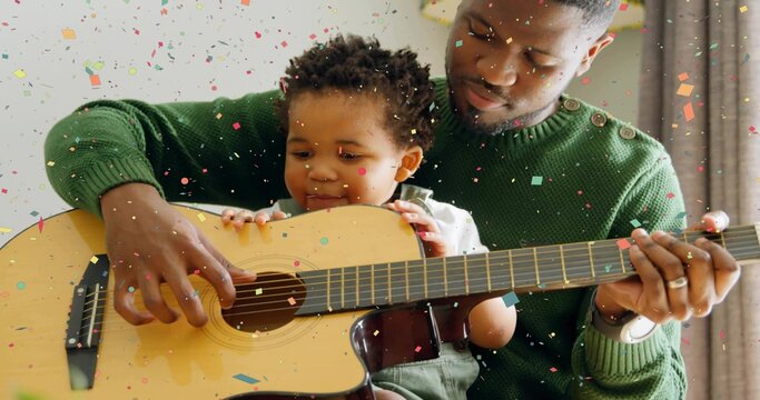 Playing father and baby guiding hands on home sofa with wood guitar, green sweater, green overalls