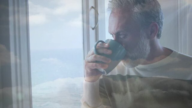 Senior man lifting teal mug sipping musing turning as moving cloud overlay covering window, health