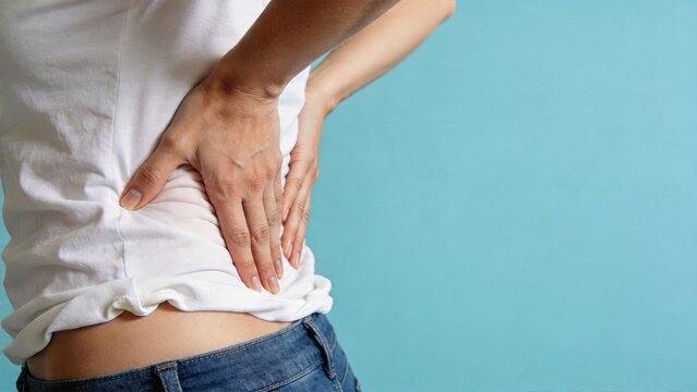 close up of woman in white shirt holding lower back in pain with hand on waist suffering from acute lumbar spine discomfort or sciatica syndrome due to muscular strain stress