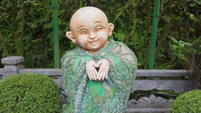A smiling stone statue of a child, adorned with moss and pebbles, sits in a lush garden in Ba Na Hills, Da Nang, Vietnam. The statue evokes a sense of peace and joy.