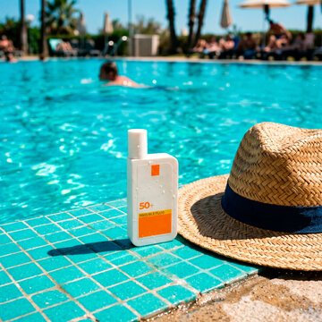 Protecci&oacute;n solar en vacaciones: Sombrero de paja y crema protectora SPF 50 junto a una piscina soleada.