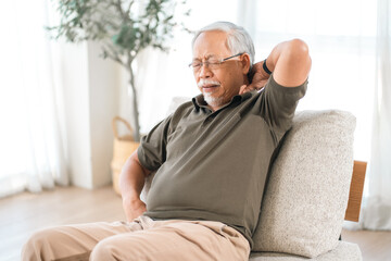 Fototapeta premium Elderly man with neck pain, sitting on sofa in bright room, discomfort and fatigue