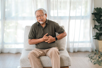 Fototapeta premium Chest pain elderly man sitting on chair at home with hand on chest and pained face, emergency discomfort, indoor daylight