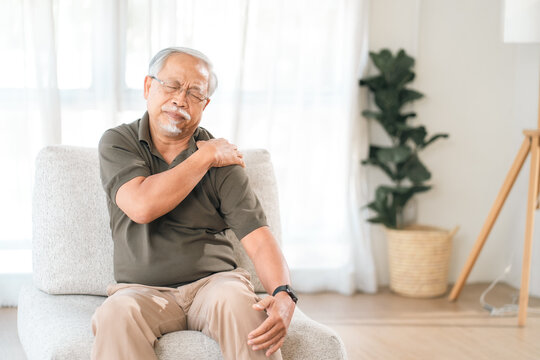 Elderly man shoulder pain sitting on chair in bright living room, painful posture and discomfort