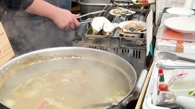 Street food chef grilling fresh oysters and scallops while cooking hot crab miso soup in large pot.