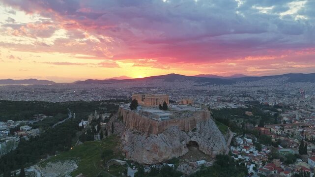Breathtaking aerial footage capturing the majestic Acropolis of Athens and the Parthenon temple, illuminated by the vibrant, colorful clouds of a stunning sunset over the cityscape