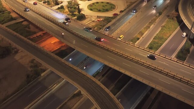 Dramatic aerial view of a multi-level highway interchange with heavy car traffic at night, showing vehicles with their headlights on driving across elevated roads and complex junctions
