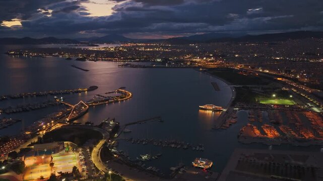 Stunning aerial perspective of the illuminated athens coastline at dusk, showcasing the bustling marinas of faliro and the vibrant city lights against a dramatic cloudy evening sky