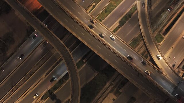 Top-down aerial view of a busy, complex highway interchange with multi-level roads and overpasses illuminated at night, showing cars driving with headlights on this urban infrastructure
