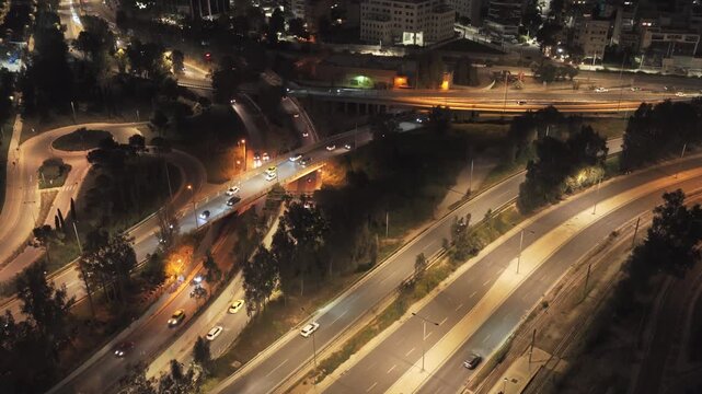 Aerial view of a complex highway interchange with busy car traffic at night, showing illuminated roads, bridges, and surrounding urban buildings creating a dynamic metropolitan cityscape