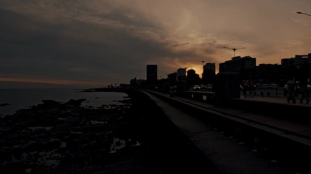 Dramatic sunset over the montevideo skyline, with cars driving along the coastal road. The dark silhouette of the city contrasts with the orange sky
