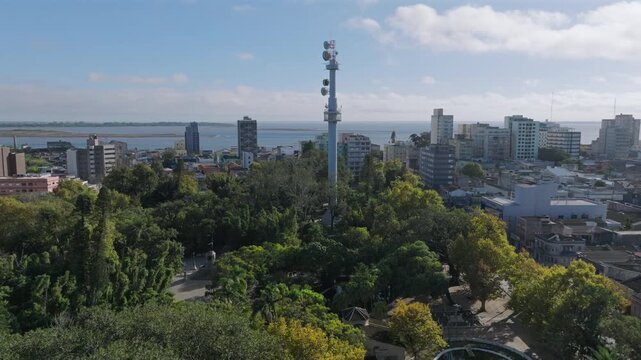 Drone advances toward tall communications tower rising above park treetops with city skyline and lagoon beyond