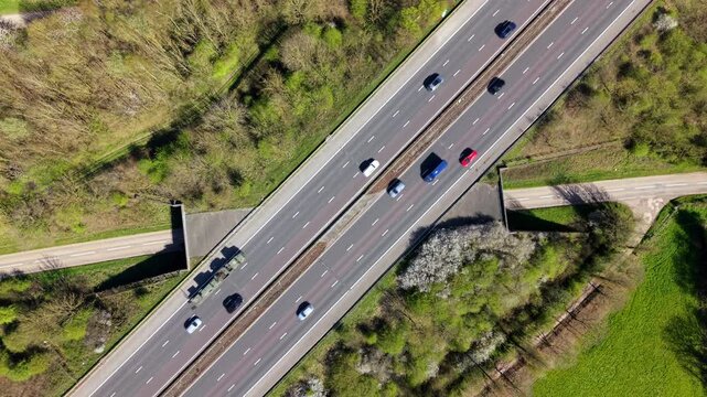 Aerial view of vehicles driving along the M1 motorway surrounded by green trees and spring blossoms in Leicestershire, England, United Kingdom.