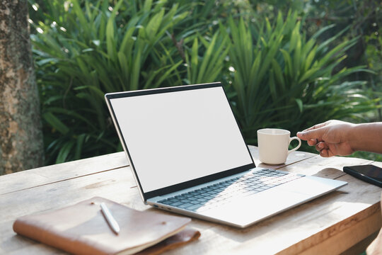 Remote work lifestyle in morning sunlight with laptop on outdoor wooden desk, hand reaching for coffee cup, balanced productivity and mindful break in natural garden workspace environment