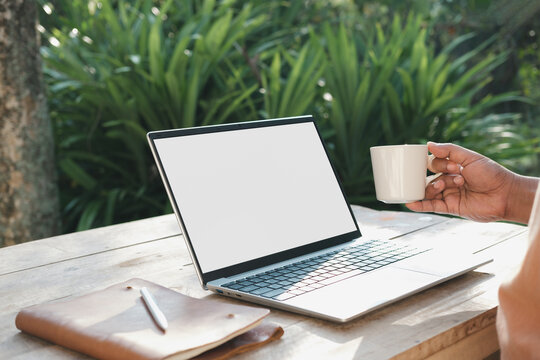 Remote work lifestyle holding coffee cup beside laptop in soft morning light, outdoor wooden workspace in garden, relaxed productivity and slow living concept with natural environment