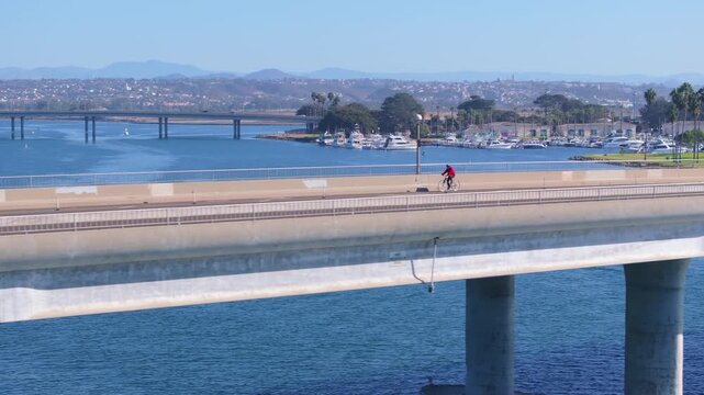 Aerial view of Mission Bay Park in San Diego featuring concrete bridge over calm blue water with cyclist in red jacket, passing cars, and pedestrians.