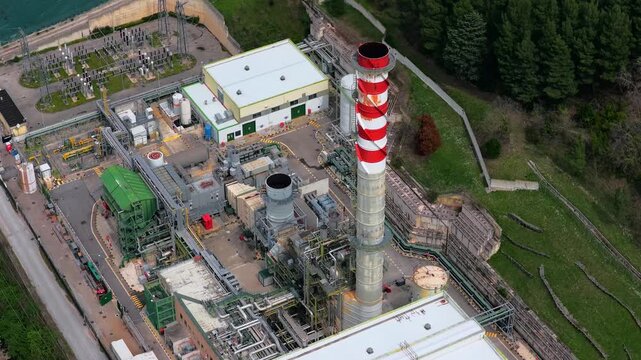 Aerial view of an industrial power plant with a red and white striped chimney and electrical substation surrounded by green hills in Bussi sul Tirino, Abruzzo, Italy.