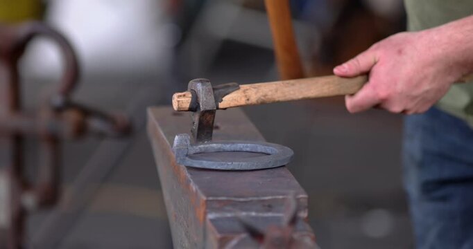 Close-up of a blacksmith using a hammer and punch to shape a metal horseshoe on a steel anvil within a traditional workshop setting.