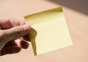 Obraz premium Hand Holding a Blank Yellow Sticky Note on a Neutral Background