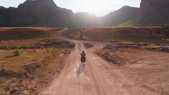 Aerial drone following motorcyclist riding on winding dusty dirt road through dramatic mountain valley at golden hour. Rustic fence posts line the rural path with stunning peaks in background.