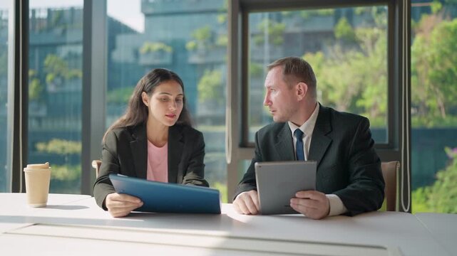 Corporate Conversation: In a modern office setting, two professionals engage in an in-depth discussion, sharing ideas and expertise as they analyze documents and utilize technology.