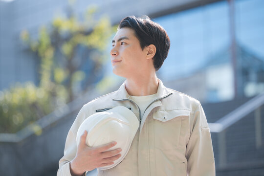 ヘルメットを持つ若手作業員のポートレート 建設現場と成長、ビル背景に未来や希望を見つめる、見上げるイメージ