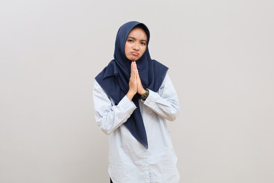 A young Asian woman in hijab needs help, has a sad and hopeful facial expression, purses her lower lip and puts her palms together, pleading or asking for help, her gaze is blank.