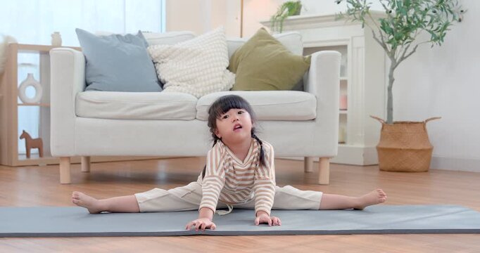 Asian preschool girl sits in wide leg stretch on exercise mat in bright living room, practicing yoga with calm expression and steady posture, building flexibility, balance, strength, wellness, poise.