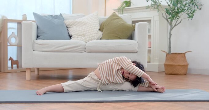 Asian preschool girl performs side stretch on exercise mat in bright living room, reaching toward foot during yoga practice with flexible control, building balance, strength, wellness, confidence.