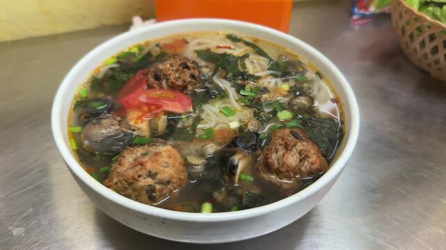 Traditional Vietnamese bun oc soup with pork meatballs, river snails, rice vermicelli, tomatoes, and fresh herbs served in Hanoi Old Quarter street food stall.