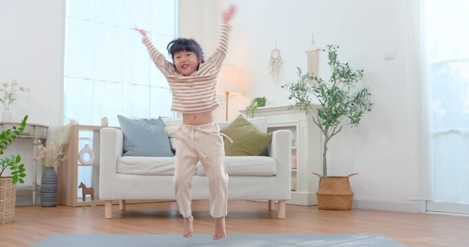 Asian preschool girl jumps above exercise mat in bright living room after yoga practice, raising arms with joyful energy and healthy movement, building strength, balance, confidence, active wellness.
