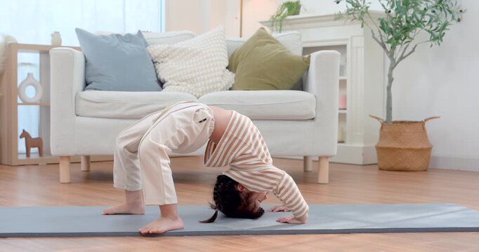 Asian preschool girl performs wheel pose on exercise mat in bright living room, arching body with strong control during yoga practice, building flexibility, balance, strength, courage, wellness.