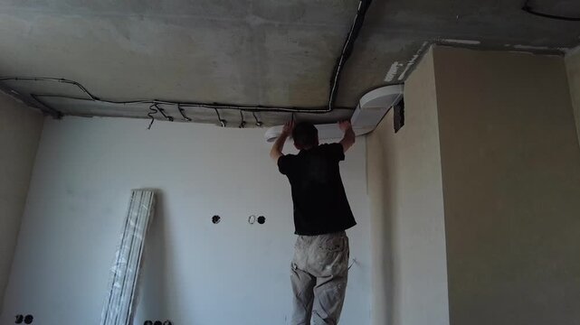 A man is renovating a room, installing a range hood in the kitchen. Unplastered walls and building materials are visible, ready for use. The work is being done during the day.