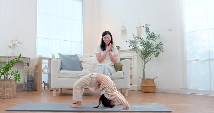 Asian preschool daughter performs bridge pose on yoga mat at home while single mother claps from sofa, encouraging confidence, flexibility, body control, playful fitness, healthy development daily