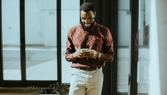 Smiling man dressed in brown shirt using smartphone in modern office space