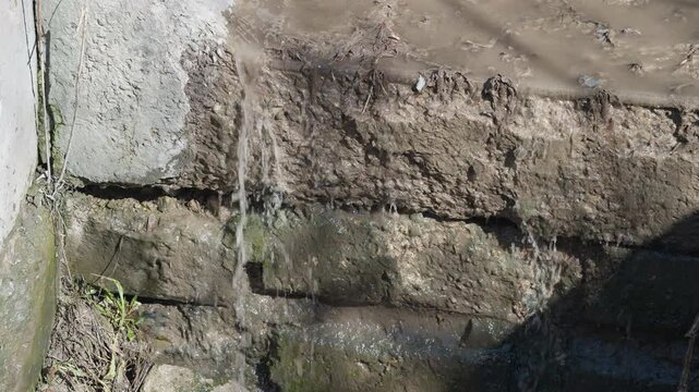 Concrete wall wastewater leak dripping from weathered curb into shadowed drain, moss and sediment streaks, slow steady flow, erosion texture, urban stormwater scene, closeup detail
