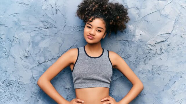 Young female teen in gray crop top with curly updo winking confident pose against textured blue wall