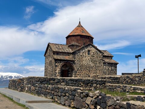 Historic Sevanavank Monastery overlooking Lake Sevan and snow-capped Caucasian mountains under a bright blue sky with white clouds, Armenia