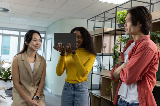 Diverse coworkers standing together sharing black tablet, leaning on desk near laptop and plants