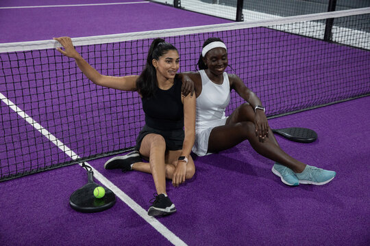 Diverse female athletes sitting on purple court in athletic wear with black paddles and green ball