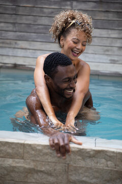 Diverse couple riding piggyback laughing against tiled pool edge at home in swimwear and sunglasses