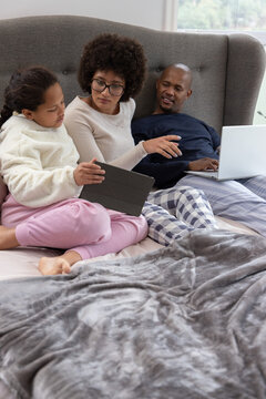 Diverse family sitting on bed, mother in sweater pointing at tablet, father in pajamas using laptop