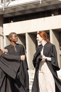 Diverse female graduates standing and chatting on campus plaza wearing black gowns holding cap