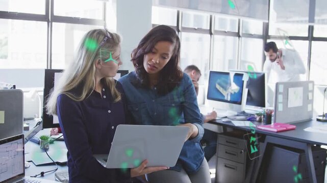 Design team reviewing laptop, dark-haired pointing and tapping, green icons showing over desks