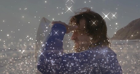 Fototapeta premium Facing surfer holding white surfboard on wet sand shading eyes wearing blue wetsuit sparkle overlay
