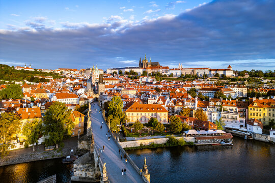 Morning light casts a soft glow over Prague Castle and Charles Bridge. The Vltava River reflects the historic buildings and vibrant rooftops of the old town, creating a serene atmosphere.